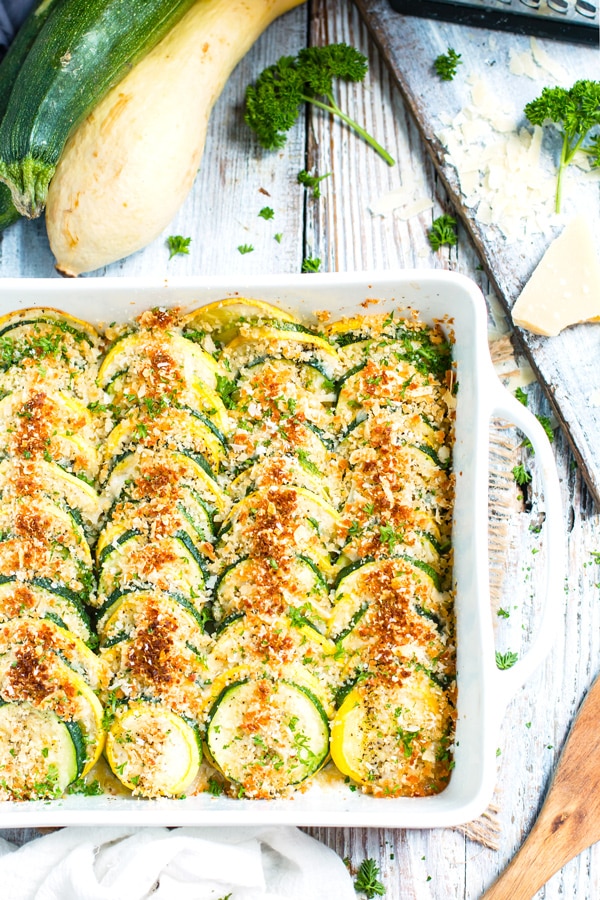 A white baking dish full of healthy squash casserole with a zucchini and yellow squash next to it.
