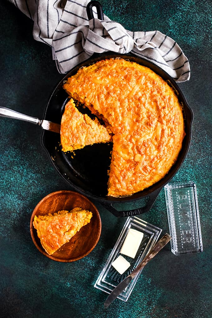 A skillet of Pumpkin Cream Cheese Skillet Cornbread with two wedges cut and one on a wooden plate and the skillet sitting on a speckled green background.